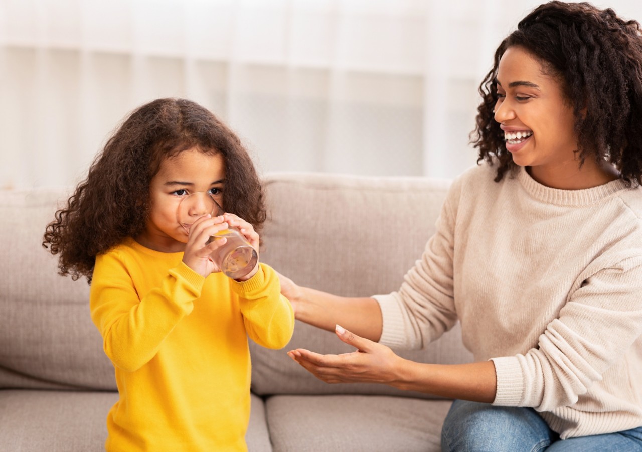 母亲的关心。快乐的非裔美国妇女给坐在家里沙发上的小女儿喂水。一些:1199674829