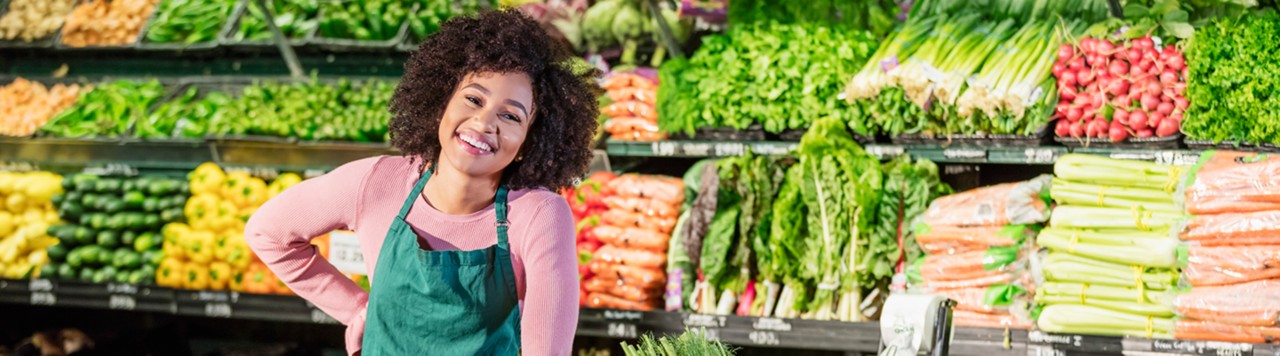 一位20多岁的年轻非裔美国女性在杂货店的农产品货架上工作，周围摆满了新鲜的水果和蔬菜。