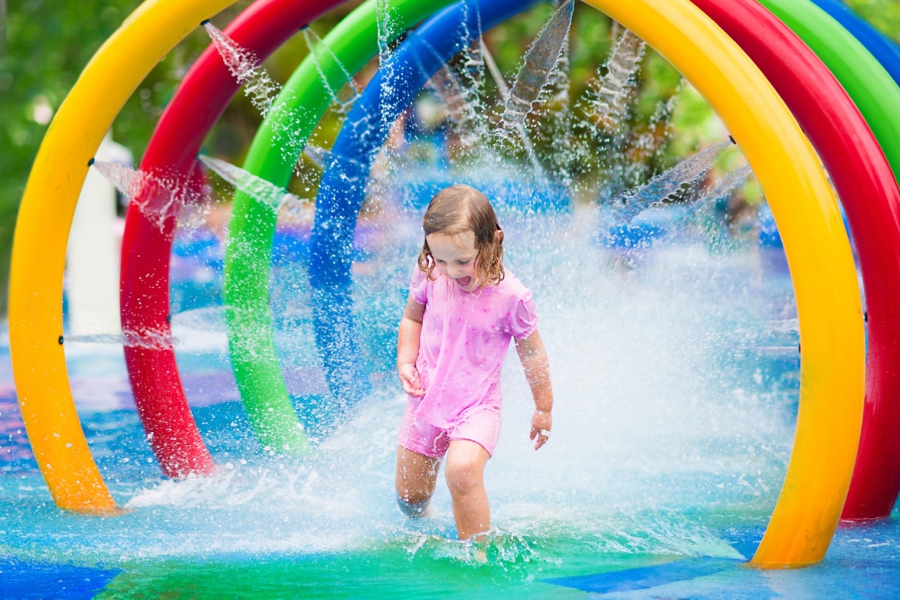 快乐的小蹒跚学步的女孩通过一个充气玩具洒水车有乐趣的水飞溅在游泳池享受一日游到水上游乐园在夏季家庭度假＂class=