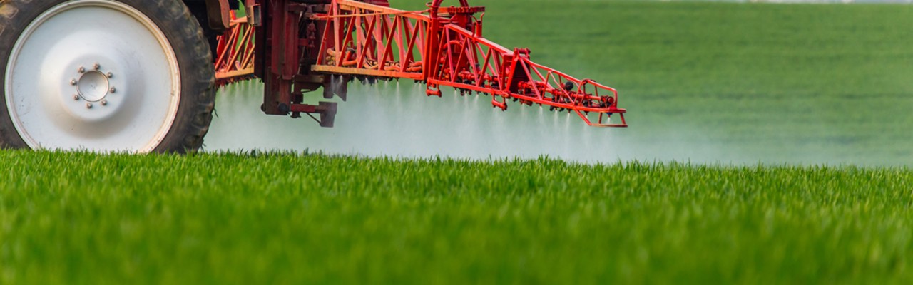 一辆红色拖拉机向一片绿色作物喷洒农药的裁剪图片。＂title=